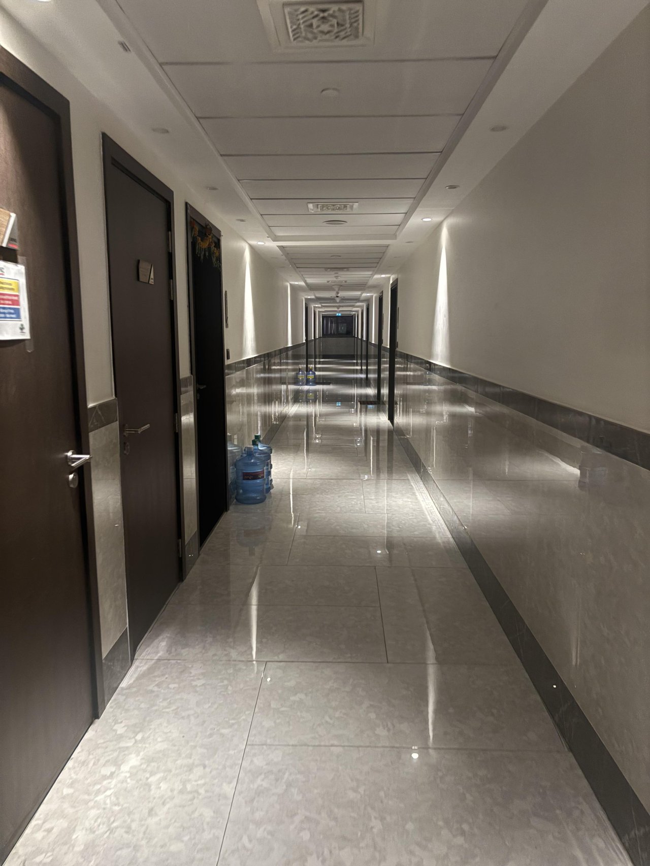 A long empty hallway with dark doors, glossy tiles, reflective wall panels, and water bottles.