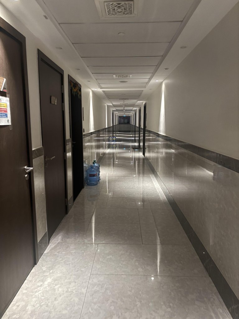 A long empty hallway with dark doors, glossy tiles, reflective wall panels, and water bottles.