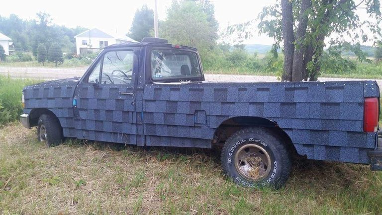 A parked pickup truck has blue asphalt roofing shingles attached across its hood, doors, and bed.